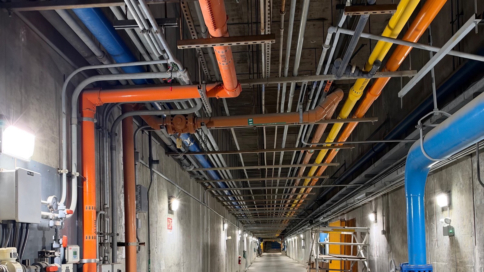 A long concrete gallery lined with colour-coded chemical feed and water pipes inside a treatment plant.